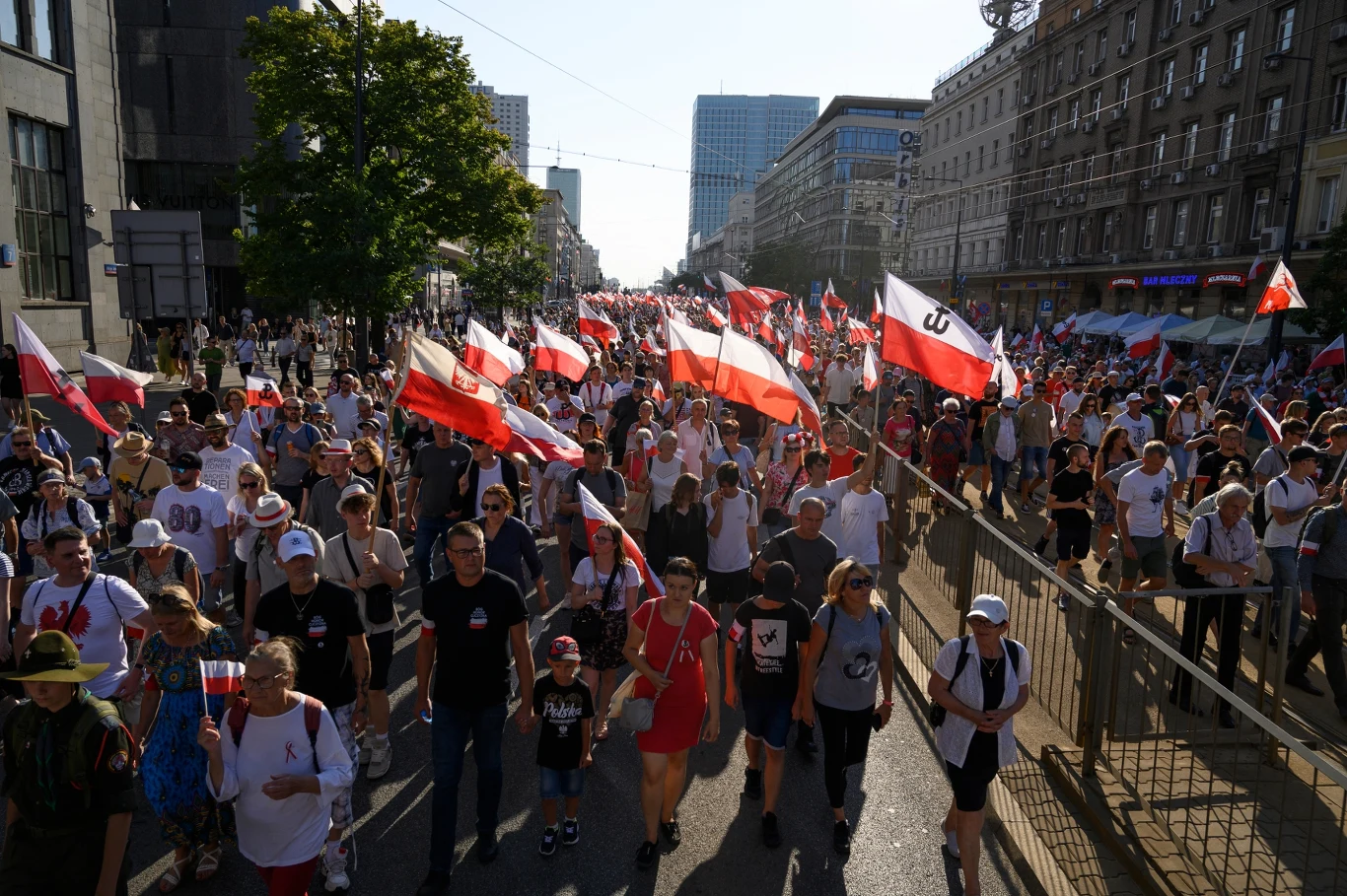 Obchody rocznicy Powstania Warszawskiego Tłum ludzi maszeruje ulicą miasta, większość trzyma w rękach biało-czerwone flagi Polski, niektórzy mają na sobie ubrania w barwach narodowych, wokół wysokie budynki i drzewa, atmosfera pokojowa i manifestacyjna.