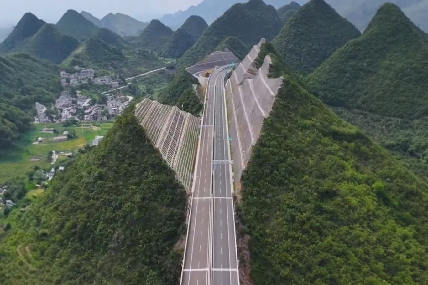 Przecięli góry na pół, aby środkiem poprowadzić autostradę. Szeroka autostrada przebiega przez przecięte wzgórza porośnięte bujną zielenią, w tle widoczne są góry oraz niewielka wieś po lewej stronie.