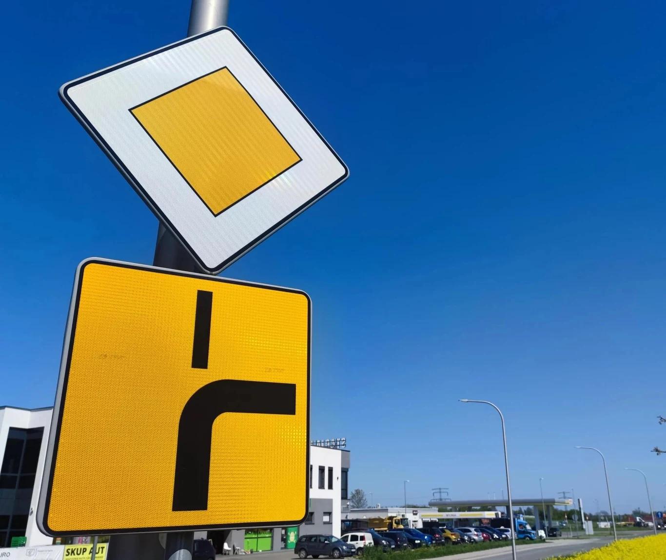 Kiedy używać kierunkowskazu na skrzyżowaniu łamanym? Dwa znaki drogowe na tle niebieskiego nieba, pierwszy informuje o drodze z pierwszeństwem przejazdu, drugi ostrzega o skrzyżowaniu z drogą podporządkowaną z prawej strony, w tle widoczne budynki i zaparkowane samochody.