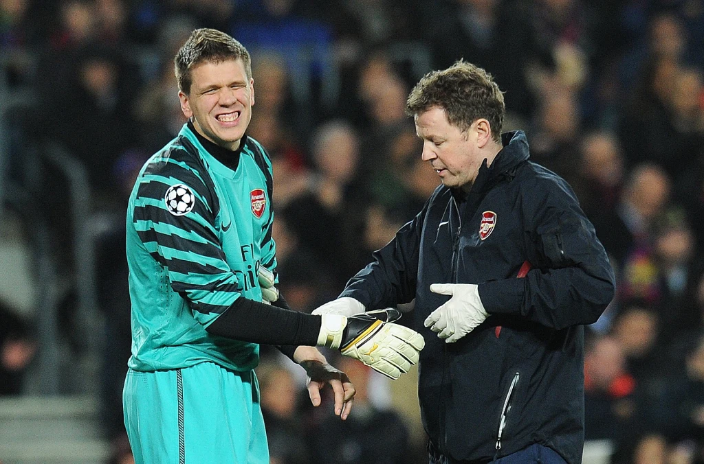 Bramkarz w turkusowym stroju piłkarskim, z logo Arsenalu na piersi, uśmiecha się podczas rozmowy z członkiem sztabu medycznego drużyny, który trzyma jego rękę i sprawia wrażenie udzielania pomocy.