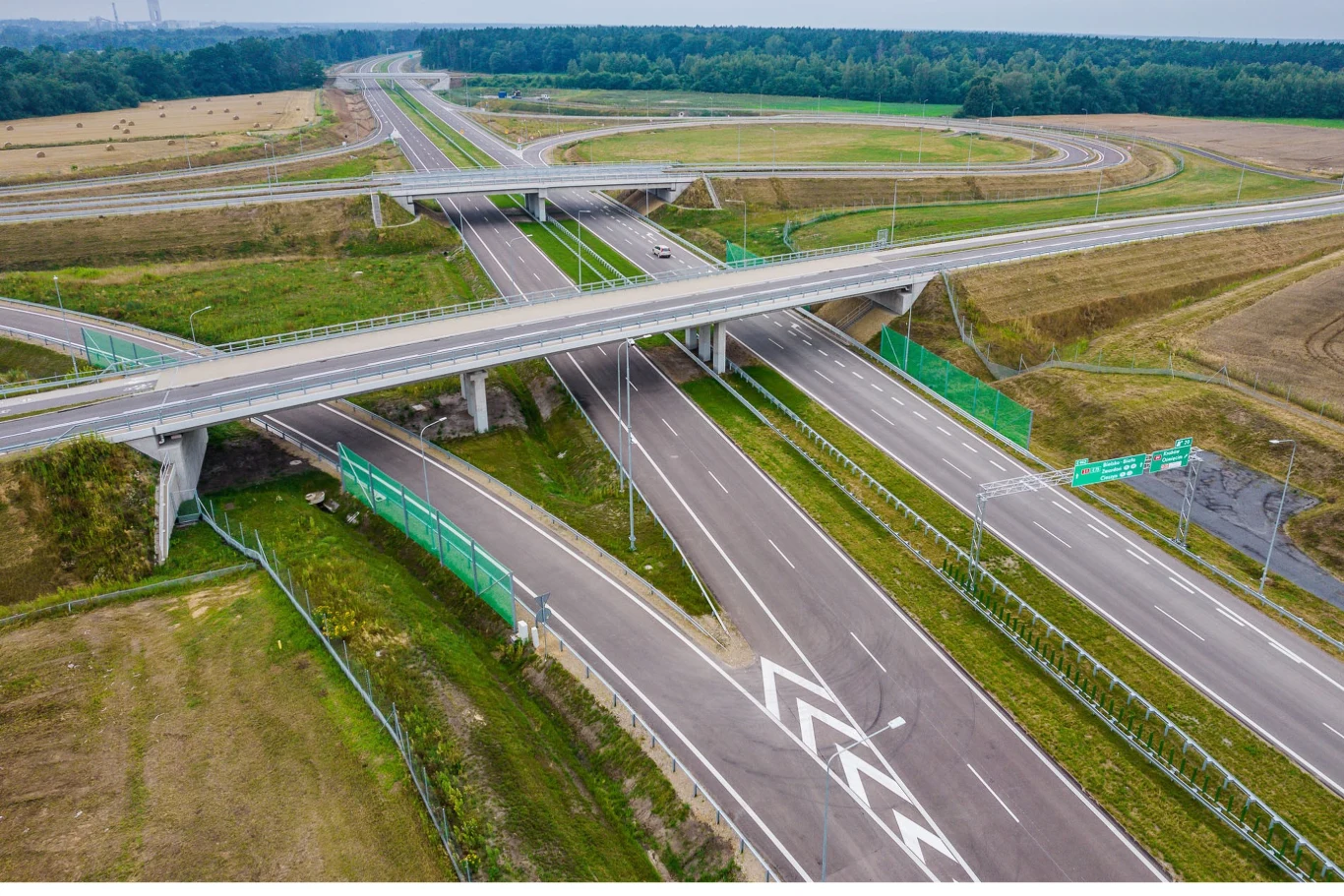 Wielopoziomowy węzeł autostradowy z kilkoma pasami ruchu, zjazdami i estakadami otoczony polami, lasem oraz ekranami dźwiękochłonnymi.