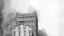 Dwóch żołnierzy siedzących za murem, obserwujących płonący budynek otoczony dymem, w tle monumentalna, zniszczona wieża.