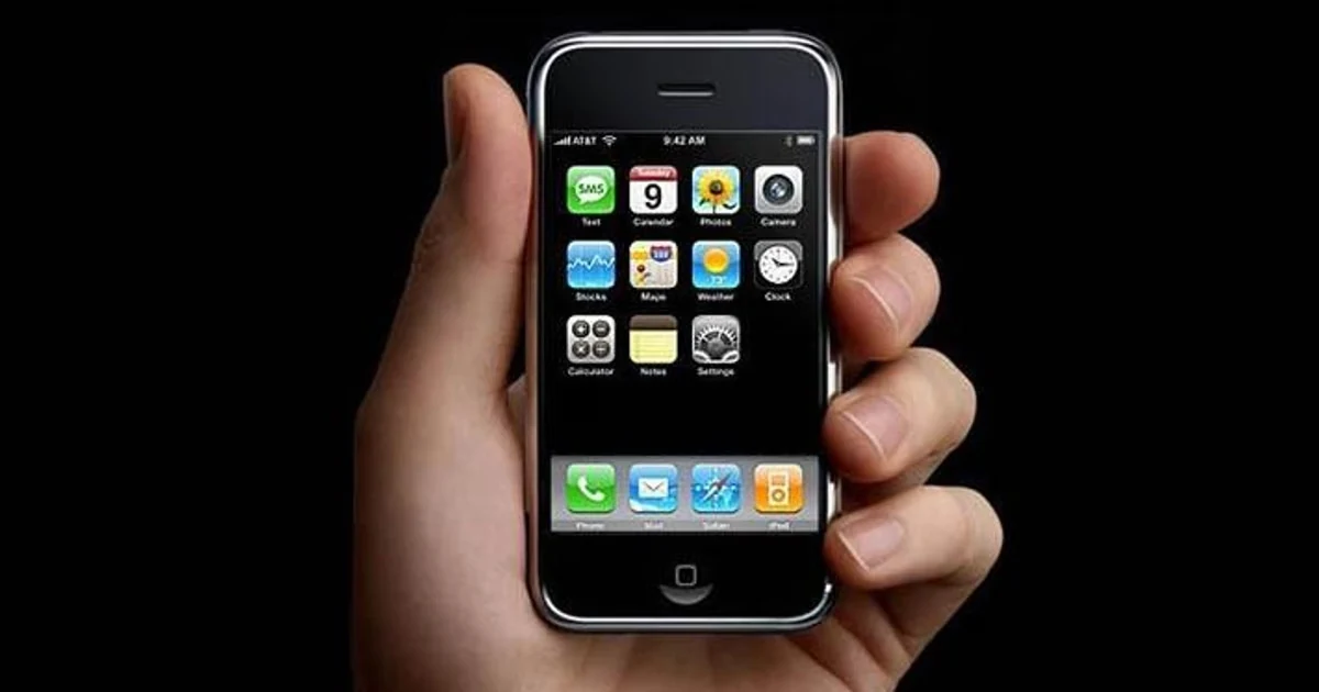 Pierwszy iPhone zaprezentowany przez Apple w 2007 r. Dłoń trzyma klasycznego smartfona z włączonym ekranem wyświetlającym podstawowe aplikacje, na czarnym tle.