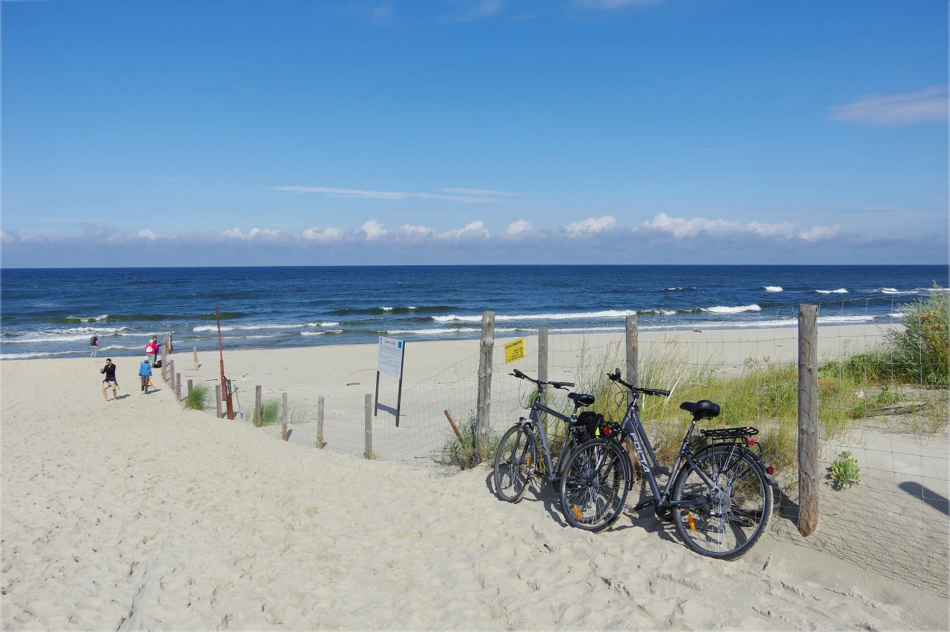 Dwa rowery stojące na piasku przy drewnianym ogrodzeniu prowadzącym na piaszczystą plażę, w tle spokojne morze i kilka osób spacerujących blisko linii wody pod błękitnym niebem.