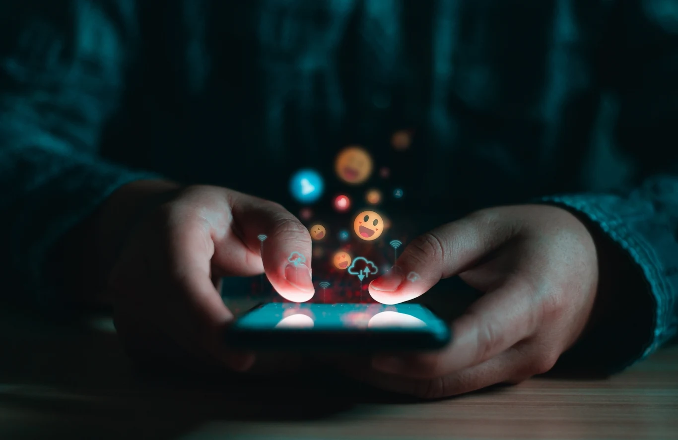 Dłonie trzymające smartfon, z ekranu unoszą się kolorowe emotikony, symbolizujące aktywność w mediach społecznościowych i komunikację cyfrową.