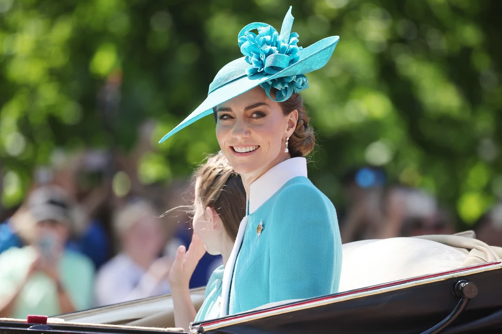Podczas tegorocznego Trooping the Colour Kate zdecydowała się na klasykę, ale w odważnym kolorze Elegancko ubrana kobieta w turkusowym kapeluszu ozdobionym kwiatami siedzi w powozie i uśmiecha się, w tle zgromadzony tłum i rozmyte drzewa.