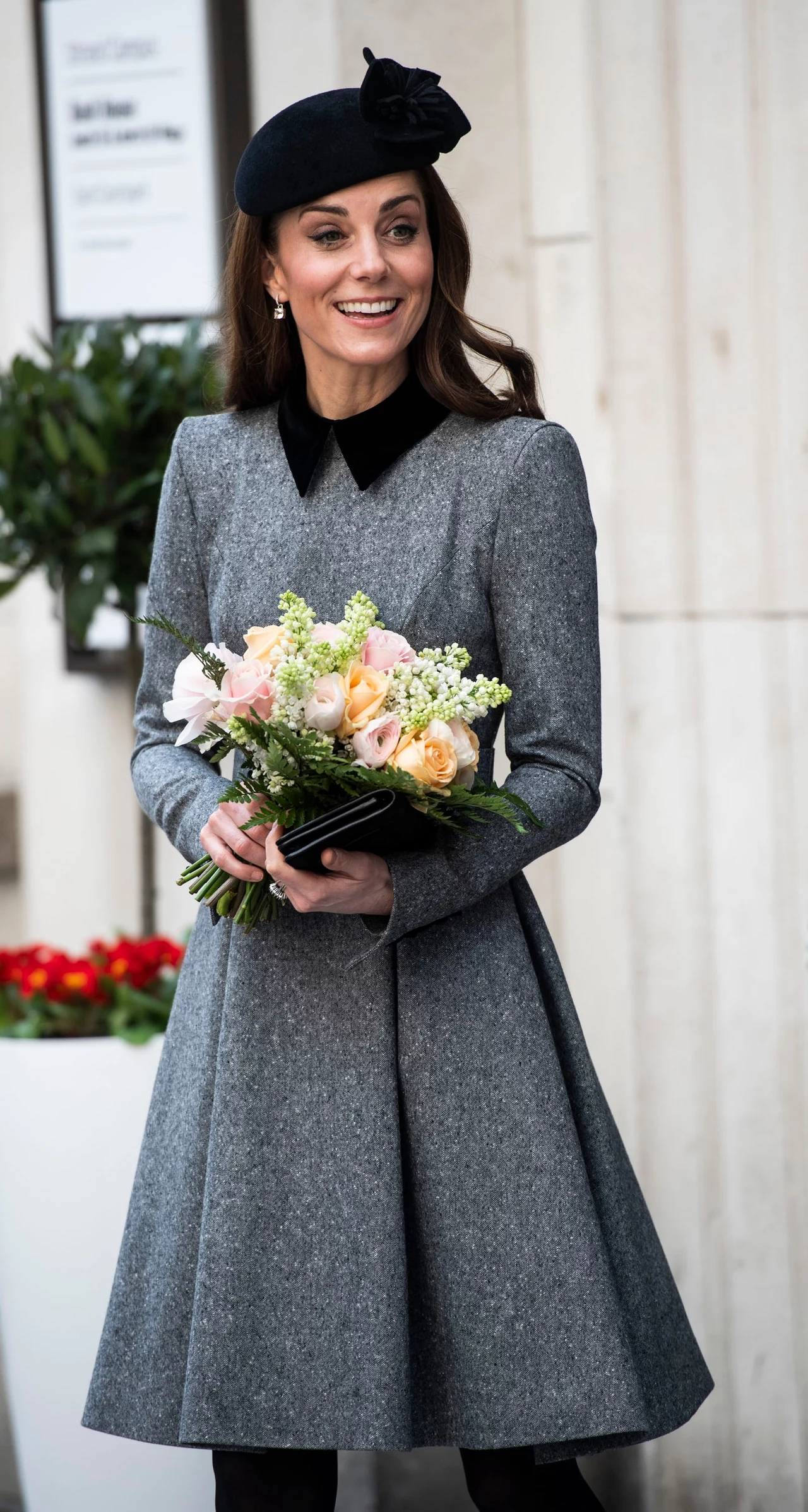 Kate z biegiem czasu wybierała coraz prostsze wzory toczków Elegancko ubrana kobieta w szarym płaszczu z czarnym kołnierzem i ciemnym beretem, trzymająca w ręku bukiet kwiatów w pastelowych kolorach, delikatnie uśmiechnięta, stoi na tle jasnej ściany z rośliną doniczkową w tle.