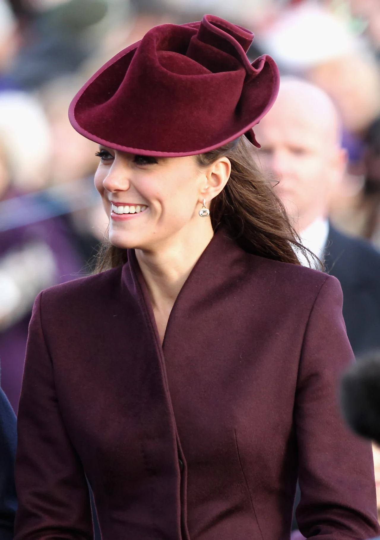 Niegdyś Kate przesadzała z rozmiarem kapeluszy Uśmiechnięta kobieta w eleganckim, bordowym płaszczu i kapeluszu z szerokim rondem o kształcie przypominającym kwiat, stojąca na tle rozmytego tłumu.