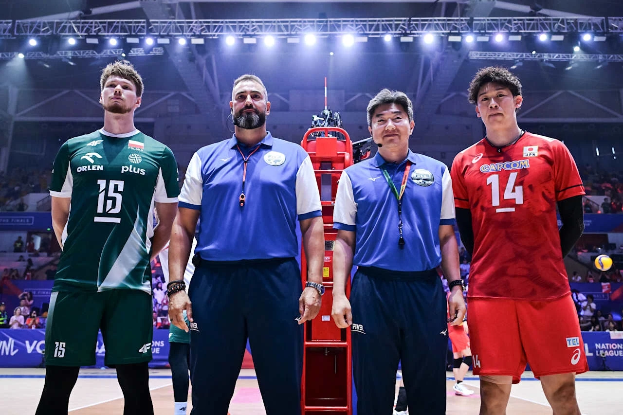Jakub Kochanowski (z lewej) jest w finałach Ligi Narodów kapitanem reprezentacji Polski Czterech mężczyzn stoi na boisku siatkarskim, dwóch z nich ubranych w sportowe stroje drużyn siatkarskich, a dwóch w niebieskich koszulkach sędziowskich, w tle widoczne trybuny i publiczność, na podłodze piłka do siatkówki.