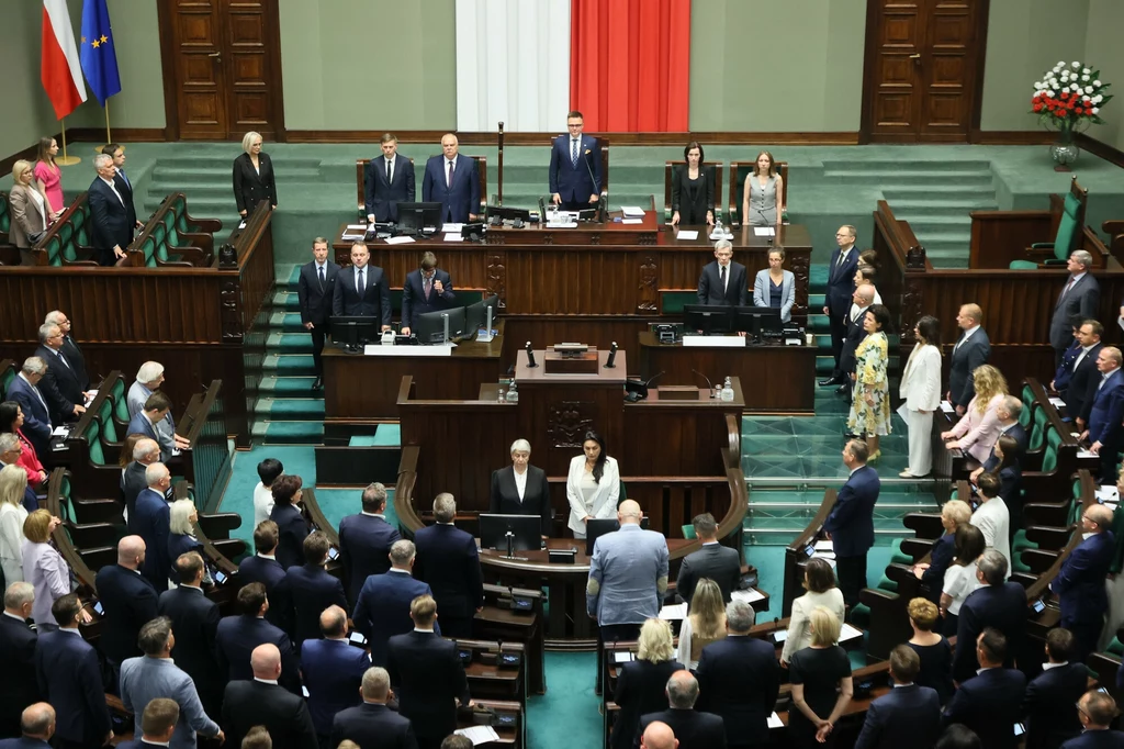 Szymon Hołownia w czasie obrad Sejmu Sala plenarna polskiego sejmu w trakcie oficjalnych obrad, posłowie stoją przy swoich miejscach, widoczna trybuna marszałka oraz flagi Polski i Unii Europejskiej