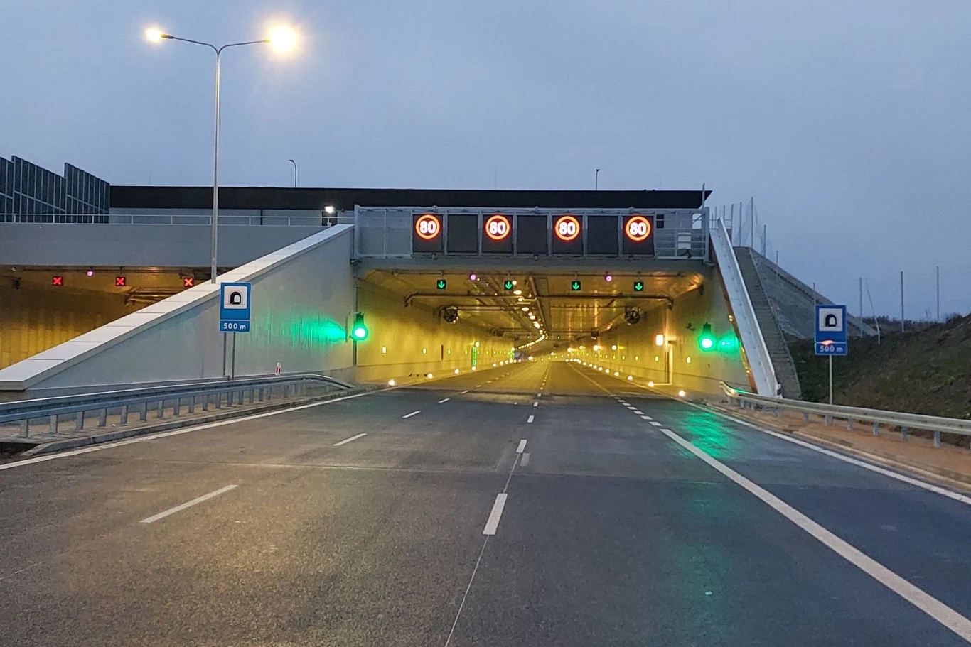 Tunel na S52 zostanie wyposażony w odcinkowy pomiar prędkości. Wjazd do nowoczesnego tunelu drogowego oznaczonego wyświetlaczami z ograniczeniami prędkości oraz zielonymi światłami na pasach wjazdowych, otoczony barierkami i znakami informacyjnymi.