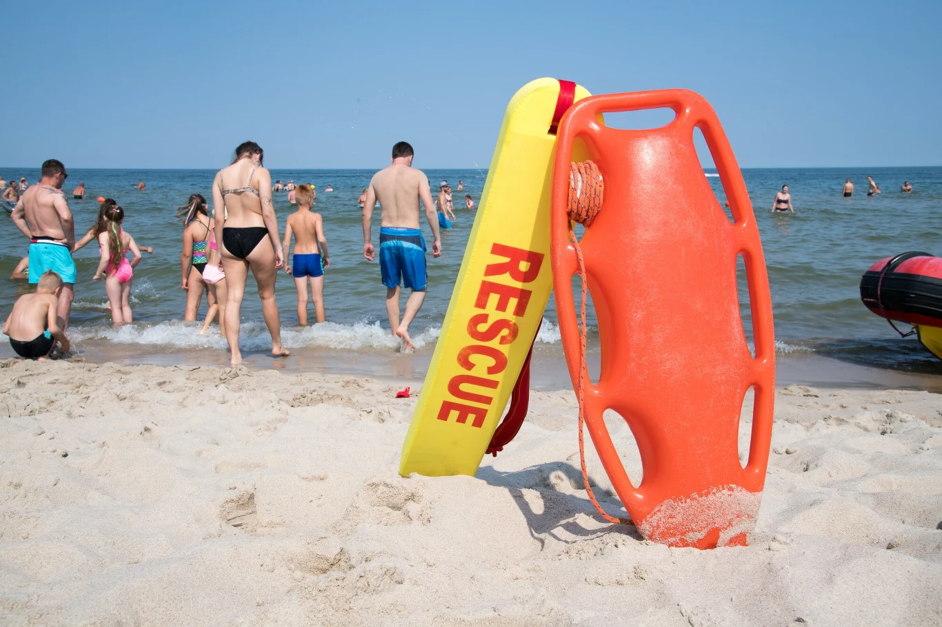 Czy Polacy wiedzą, jak wezwać pomoc nad morzem albo w górach? Plaża z grupą ludzi kąpiących się i spacerujących po piasku, w centralnej części kadru ratownicze sprzęty plastikowe: pomarańczowa deska ratunkowa oraz żółta boja z napisem RESCUE, w tle spokojne morze i błękitne niebo.