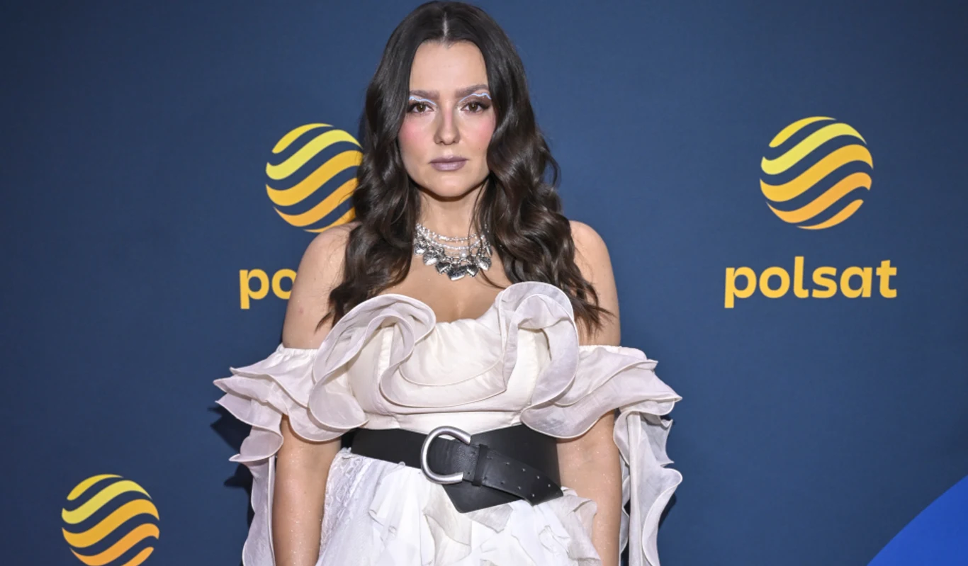 Lanberry Kobieta o długich, ciemnych włosach ubrana w efektowną, białą suknię z falbanami i szerokim, czarnym paskiem, stojąca na tle niebieskiej ściany z logo Polsatu.