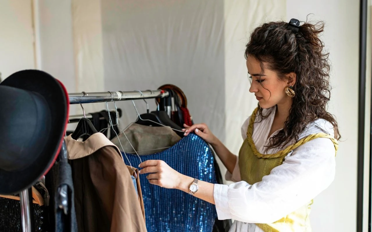 Moda z drugiej ręki? One nie wydają fortuny na ubrania Kobieta wybierająca ubrania z wieszaka, skupiona na lśniącej, niebieskiej sukience, wokół inne elementy garderoby i czarny kapelusz.