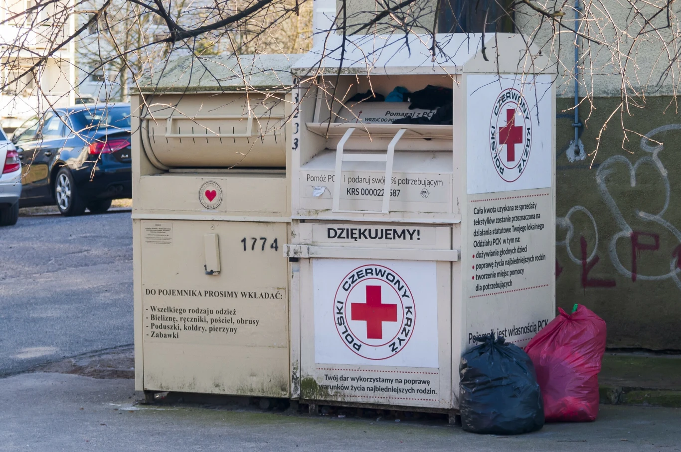 Z polskich miast znikną charakterystyczne kontenery PCK Kontener do zbiórki odzieży używanej Polskiego Czerwonego Krzyża ustawiony przy ulicy, obok dwa worki z ubraniami, w tle fragment zaparkowanych samochodów oraz ściana budynku z graffiti.