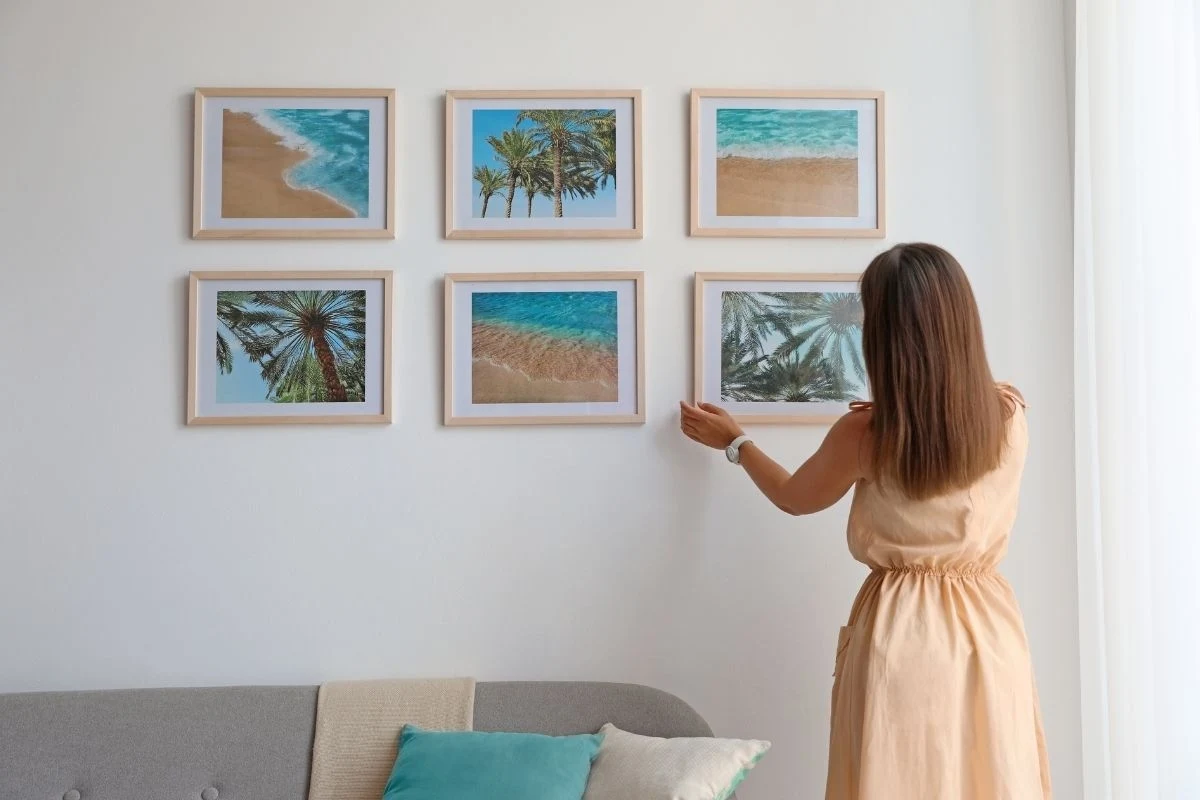 Kobieta w jasnej sukience wiesza ramkę ze zdjęciem plaży na białej ścianie, obok znajduje się pięć innych obrazów z motywami tropikalnego morza, sofa z poduszkami w odcieniach turkusu stoi na dole kadru.