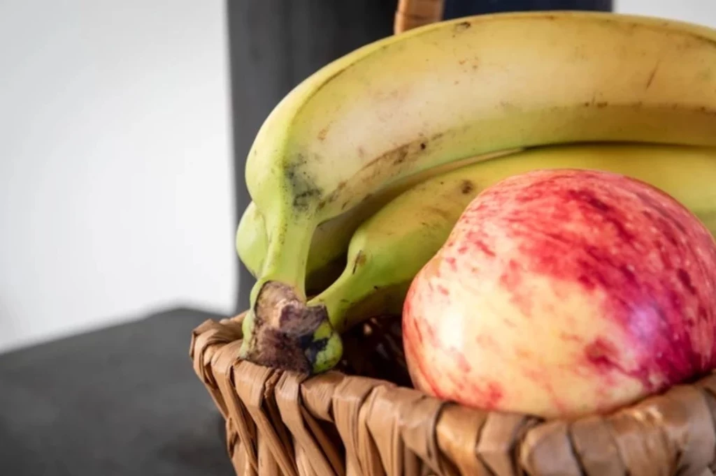 Bananów dojrzałych nie powinno się kłaść w pobliżu innych owoców klimatycznych Kosz wiklinowy z dwoma dojrzałymi bananami i czerwono-żółtym jabłkiem, ustawiony na tle neutralnej powierzchni.