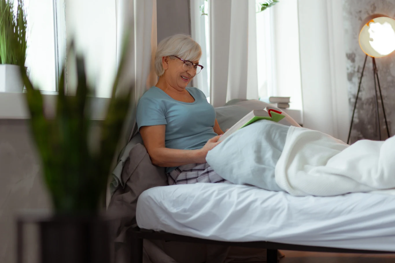 Starsza kobieta z siwymi włosami i okularami siedzi wygodnie w łóżku, uśmiecha się i czyta książkę w jasnym, przytulnym pokoju.