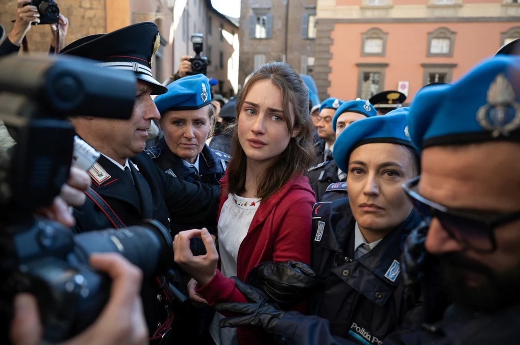 Młoda kobieta otoczona przez kilku umundurowanych funkcjonariuszy policji i straży, w tle kamienice miejskie, wokół niej obecni są także reporterzy i osoby z aparatami fotograficznymi rejestrujące sytuację.