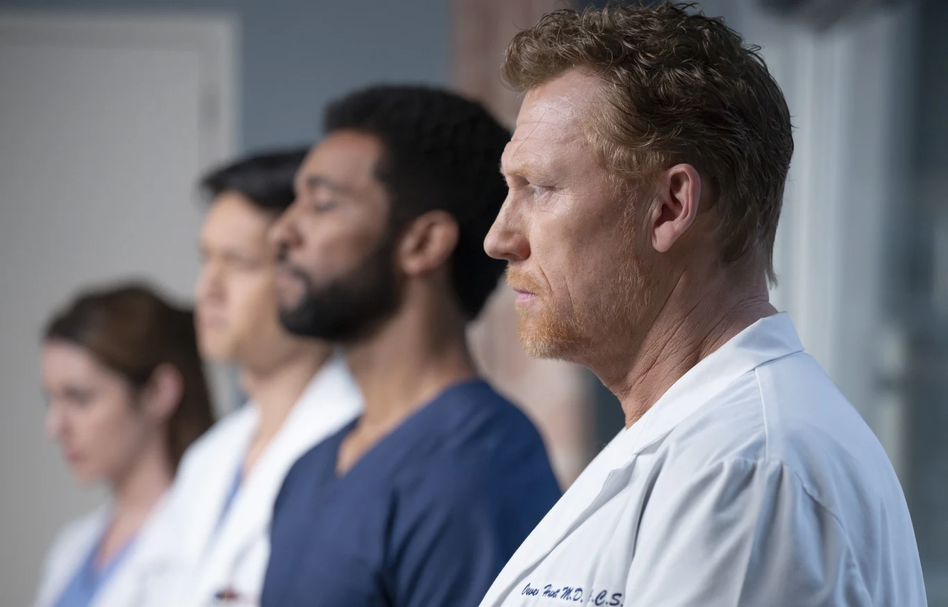 Czterech lekarzy ubranych w fartuchy medyczne stoi w szeregu, skupieni i poważni, na pierwszym planie rudowłosy mężczyzna o wyraźnym profilu, pozostali lekko rozmyci w tle.
