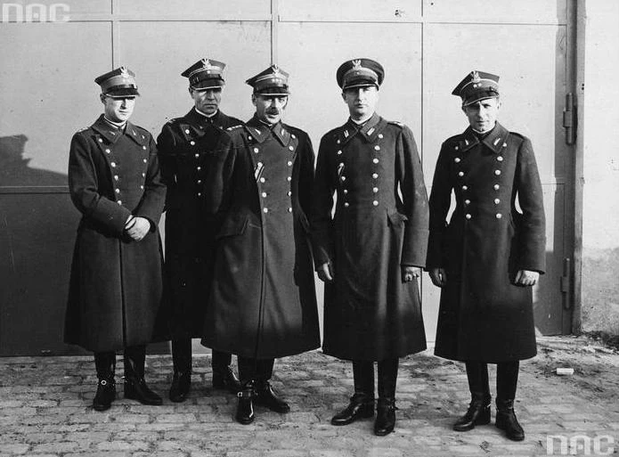 Pięciu mężczyzn w długich, wojskowych płaszczach i czapkach z daszkiem stoi w szeregu przed metalową bramą, wszyscy patrzą w stronę aparatu, tło stanowi jasny mur i brama zamknięta.