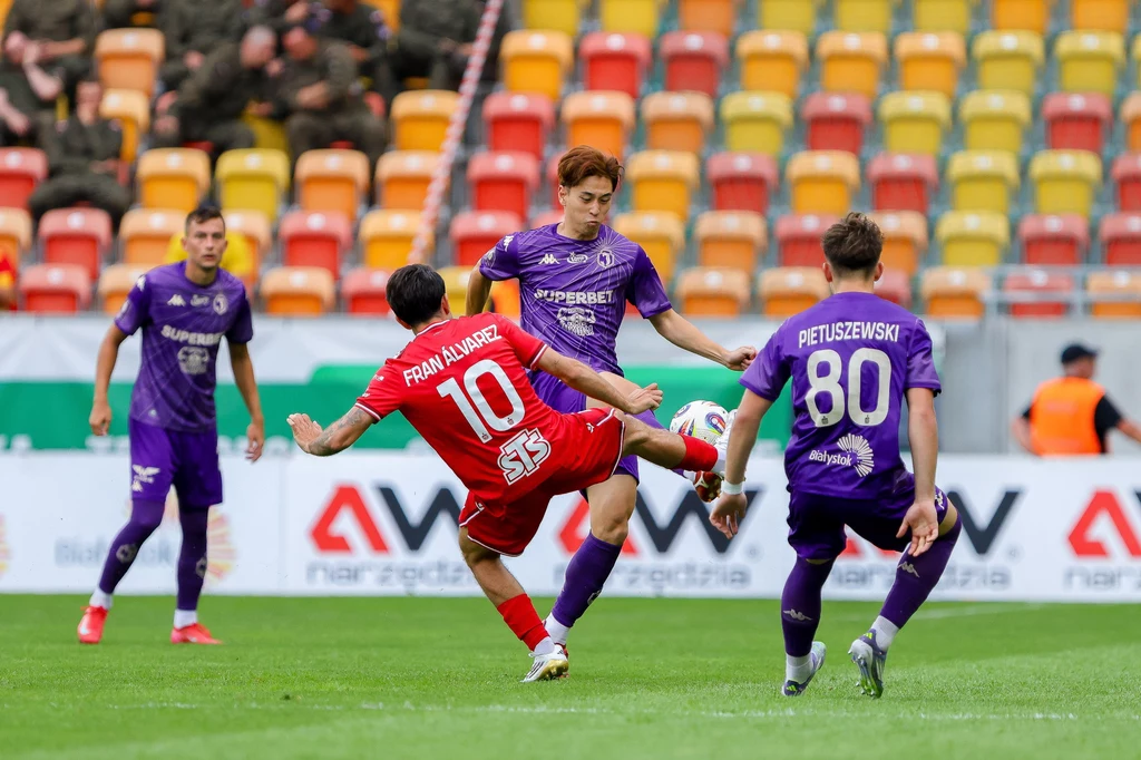 Jagiellonia Białystok - Widzew Łódź Dynamiczna akcja podczas meczu piłkarskiego, zawodnik w czerwonym stroju zagrywa piłkę, a trzech piłkarzy w fioletowych koszulkach próbuje go powstrzymać. Na trybunach widoczni są kibice i kolorowe krzesełka.