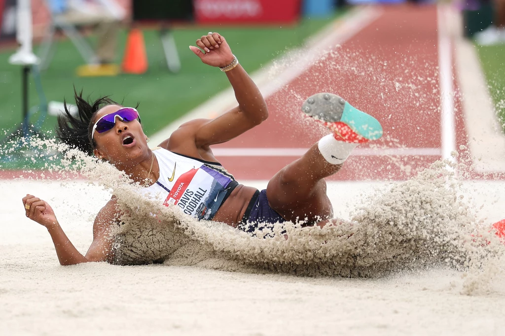 Tara Davis-Woodhall w mistrzostwach USA w Eugene Lekkoatletka ląduje w piaskownicy po skoku w dal, intensywnie rozsypując piasek, na sobie ma strój sportowy i okulary przeciwsłoneczne, tło rozmyte z elementami stadionu.