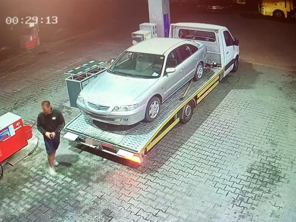 Złodziej usunął zalegające auto, a potem je zezłomował. Mężczyzna w ciemnej odzieży przechodzi obok lawety z załadowanym srebrnym samochodem osobowym, zaparkowanej przy stacji paliw oświetlonej sztucznym światłem.