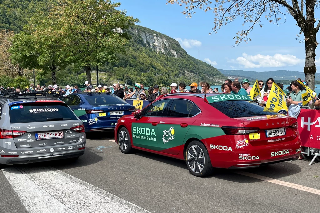 Pierwszymi autami Skody, które pojawiły się na Tour de France były modele Superb. Także dziś stanowią one trzon techniczny wyścigu. Kolorowe samochody z logo Skody i Tour de France zaparkowane na ulicy, wokół grupa kibiców trzymających żółte flagi i banery, w tle zielone drzewa oraz malownicze wzgórze.