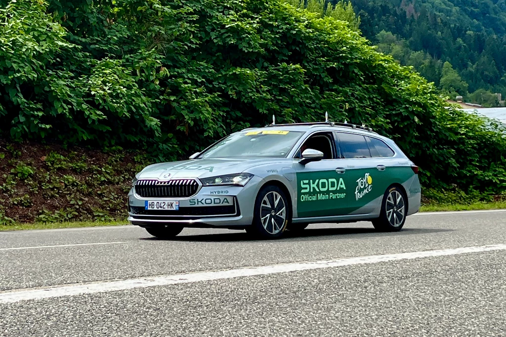 Tour de France to ekstremalnie ciężki sprawdzian nie tylko dla kolarzy. Samochód osobowy marki Škoda z oficjalnym oznaczeniem Tour de France, zaparkowany przy drodze na tle zielonych krzewów i górskiego krajobrazu.