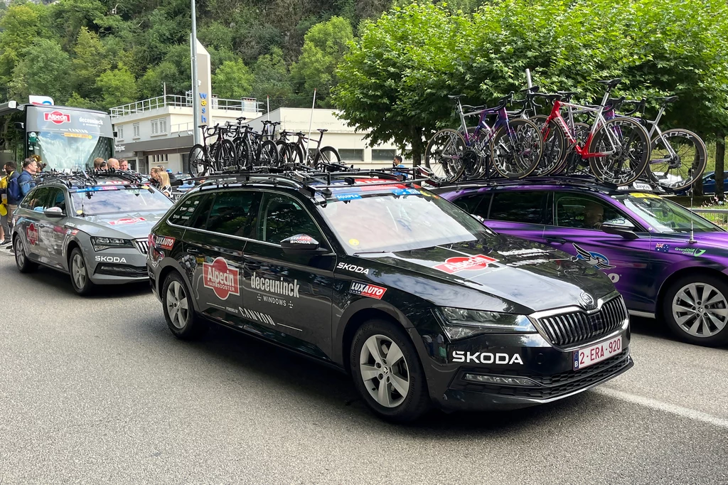 Każda drużyna dysponuje dwoma samochodami. Wiele z nich to także Skody Czarne samochody serwisowe z logotypami sponsorów, z rowerami zamontowanymi na dachach, stoją zaparkowane na ulicy, w tle widoczny autobus zespołu kolarskiego oraz drzewa.