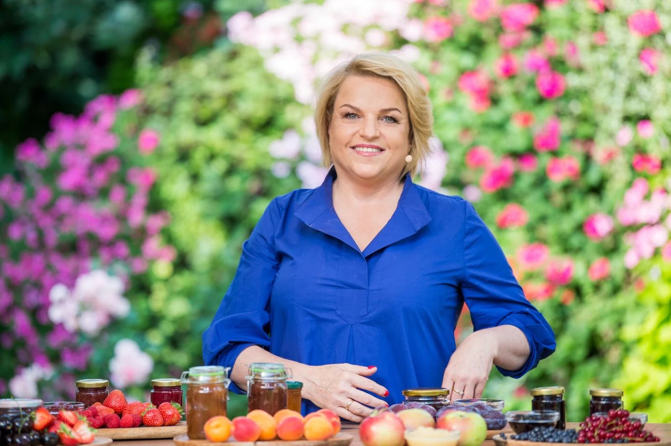 Katarzyna Bosacka przedstawiła świetny produkt na upały. Nie tylko nawadnia, ale również wspomaga pracę jelit Uśmiechnięta kobieta w niebieskiej bluzce stoi na tle ogrodu z bujną, kwitnącą roślinnością, przed nią na stole znajdują się słoiki z przetworami, świeże owoce takie jak jabłka, morele, porzeczki oraz truskawki.