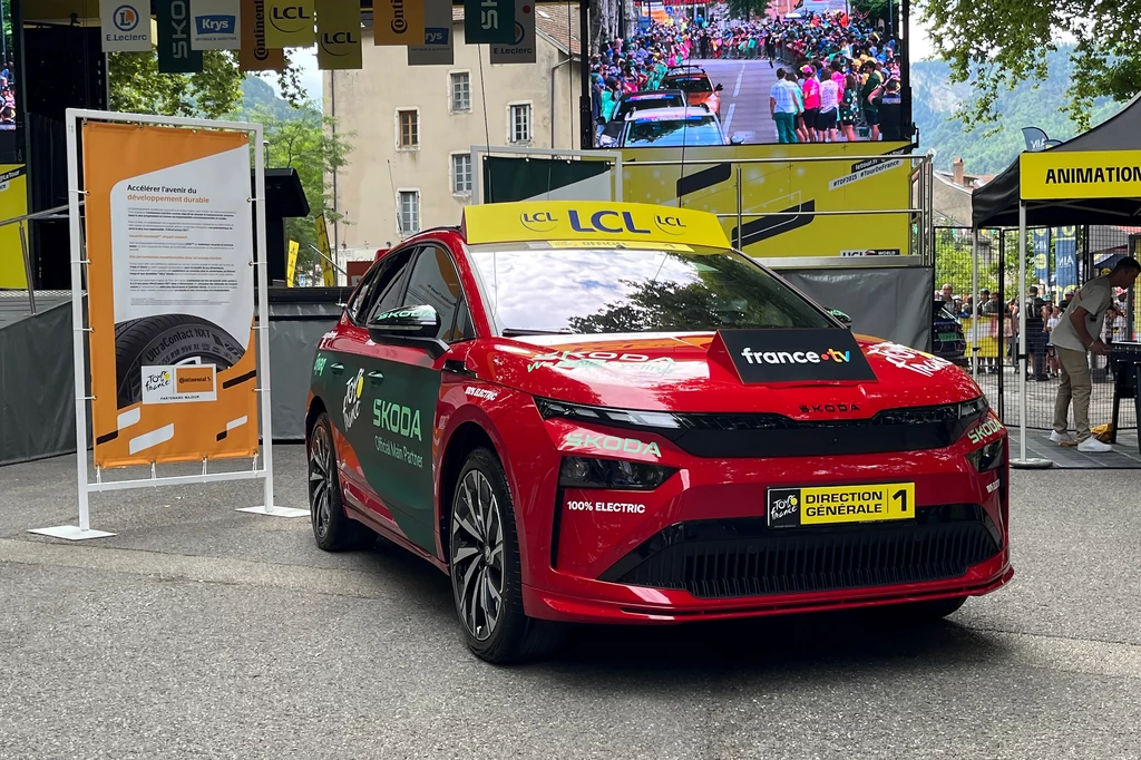 Elektryczna Skoda Enyaq - to w niej zasiada dyrektor wyścigu - Christian Prudhomme. Czerwony samochód elektryczny Škoda Enyaq z napisem 'Direction Générale' i oznaczeniami Tour de France stoi zaparkowany na ulicy przy punktach informacyjnych i banerach imprezy.