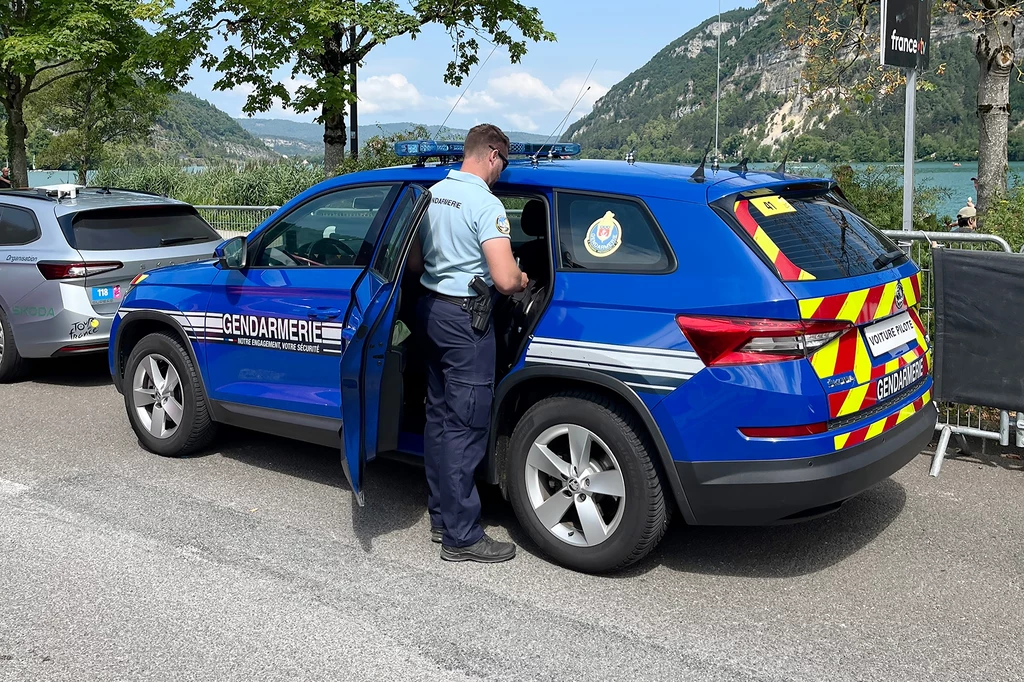 Z oferty czeskiej marki korzystają nie tylko przedstawiciele Tour de France. Policjant w mundurze stoi obok niebieskiego radiowozu żandarmerii, otwartych drzwi, w tle samochody, przyroda i góry nad jeziorem.