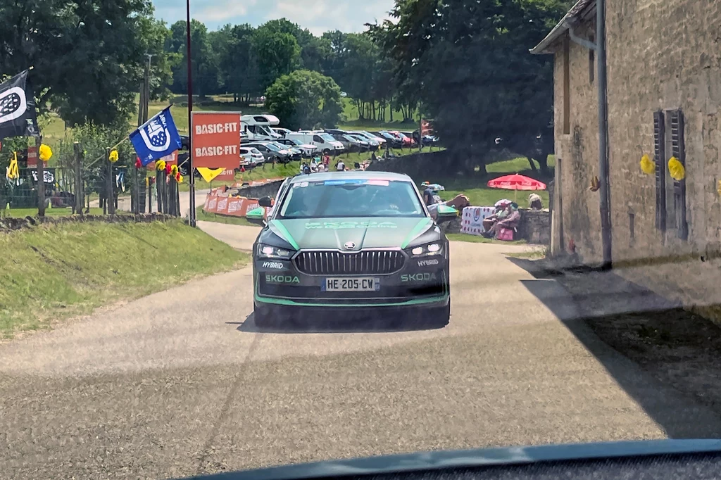 Samochody na Tour de France to warsztaty, stołówki i centra komunikacyjne na kołach Samochód Skoda z oficjalnym oznakowaniem jedzie wąską, wiejską drogą, w tle widoczne są flagi, zaparkowane auta oraz ludzie obserwujący wydarzenie sportowe.