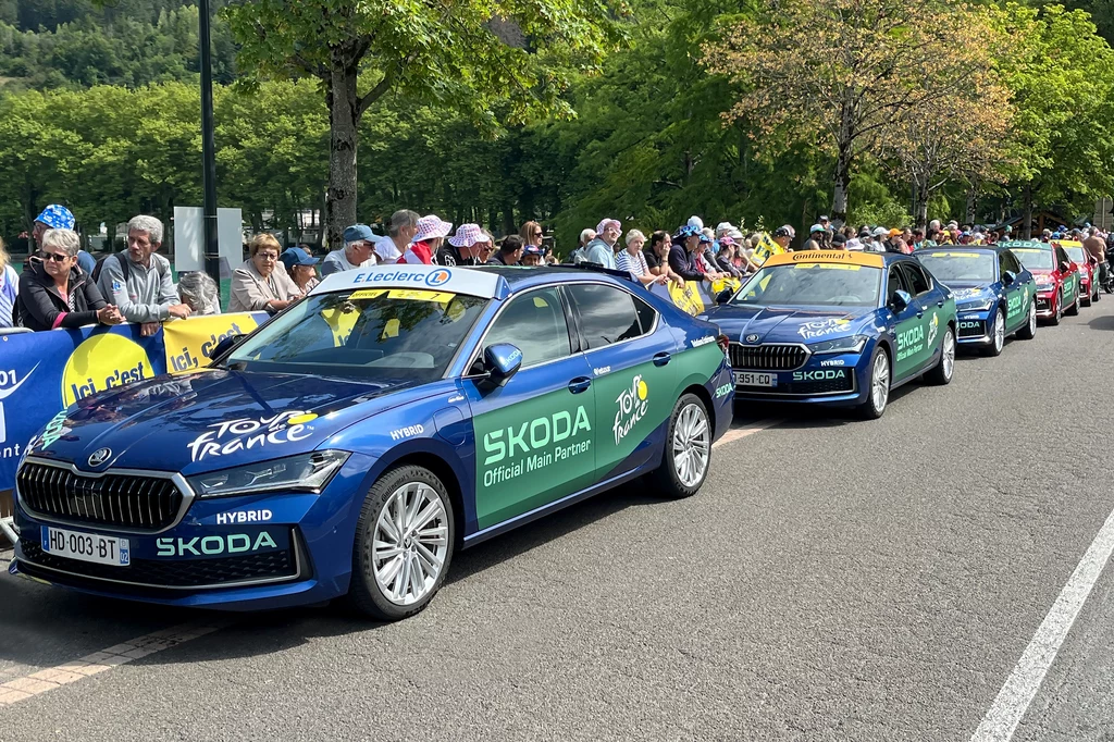W Tour de France o zwycięstwie decydują ułamki sekund. Załogi samochodów są tego świadome. Kilka samochodów marki Skoda z widocznymi logotypami sponsorów, ustawionych wzdłuż ulicy przy trasie Tour de France, tłum kibiców obserwuje wydarzenie zza barierek, po obu stronach drogi rosną drzewa.