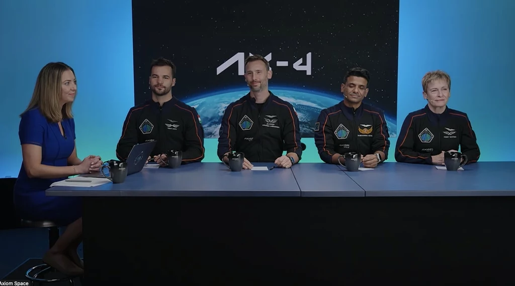 Załoga lotu Ax-4. Cztery osoby w skafandrach astronautów siedzą przy stole podczas konferencji prasowej, obok nich po lewej stronie kobieta w niebieskiej sukience prowadzi spotkanie, w tle duża grafika przedstawiająca planetę oraz napis Ax-4.