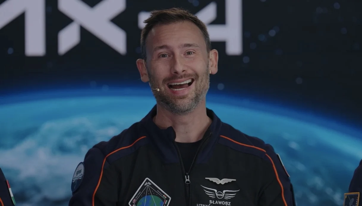 Mężczyzna w kombinezonie astronauty z naszywkami i mikrofonem na tle obrazu przedstawiającego Ziemię oraz logo SpaceX, uśmiechnięty i w trakcie wypowiedzi.