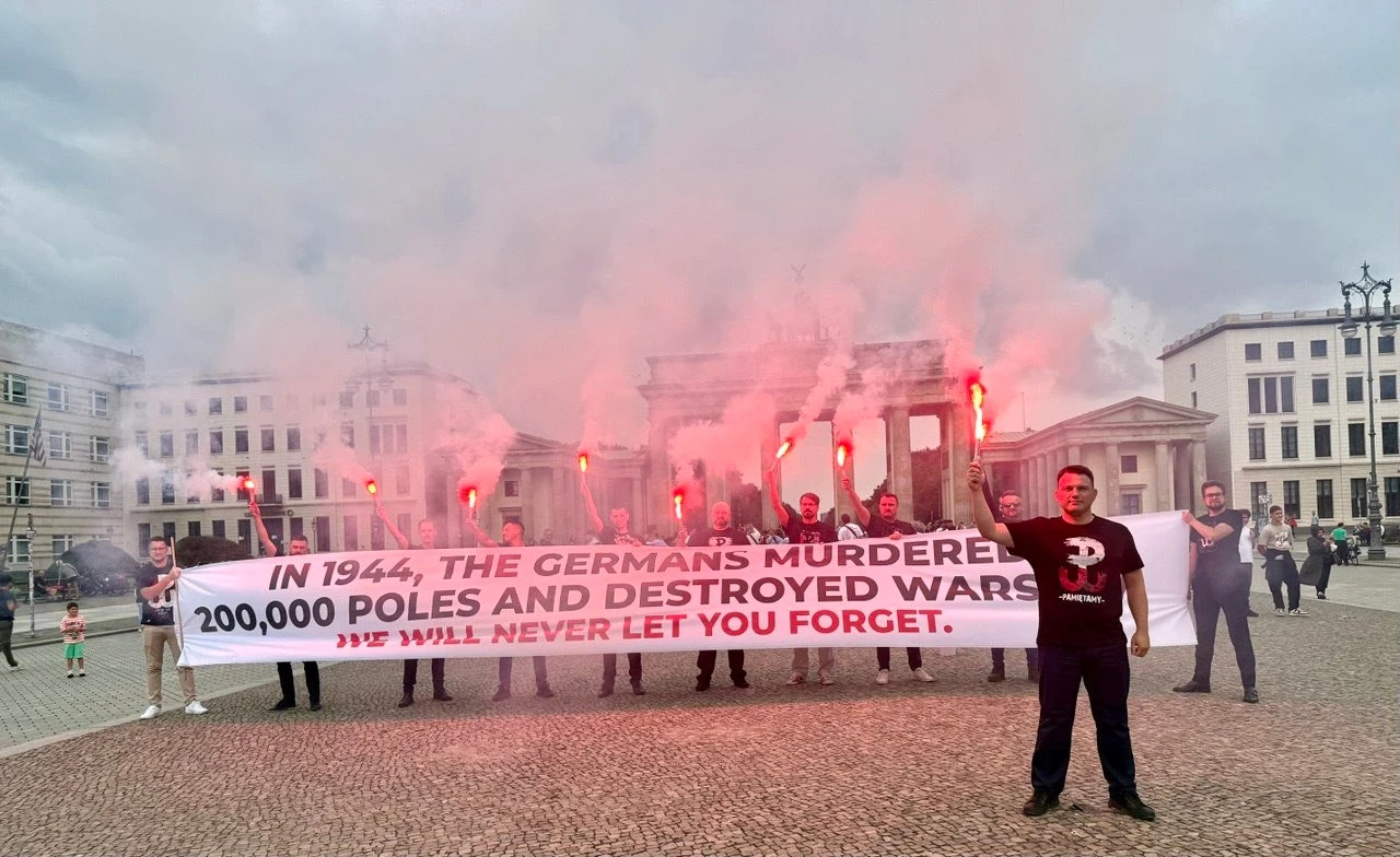 81. rocznica Powstania Warszawskiego. Sławomir Mentzen wraz z posłami pojechał do Berlina Grupa osób z zapalonymi racami trzyma duży baner z napisem dotyczącym zbrodni popełnionych przez Niemców w 1944 roku, w tle widoczna Brama Brandenburska oraz zabudowa miejskiego placu