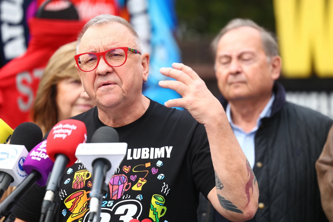 Jurek Owsiak stoi na czele organizatorów Pol'and'Rock Festival Mężczyzna w okularach z czerwonymi oprawkami i kolorową koszulką przemawia do zgromadzonych przed mikrofonami różnych mediów, za nim stoją dwie inne osoby, w tle kolorowe banery.