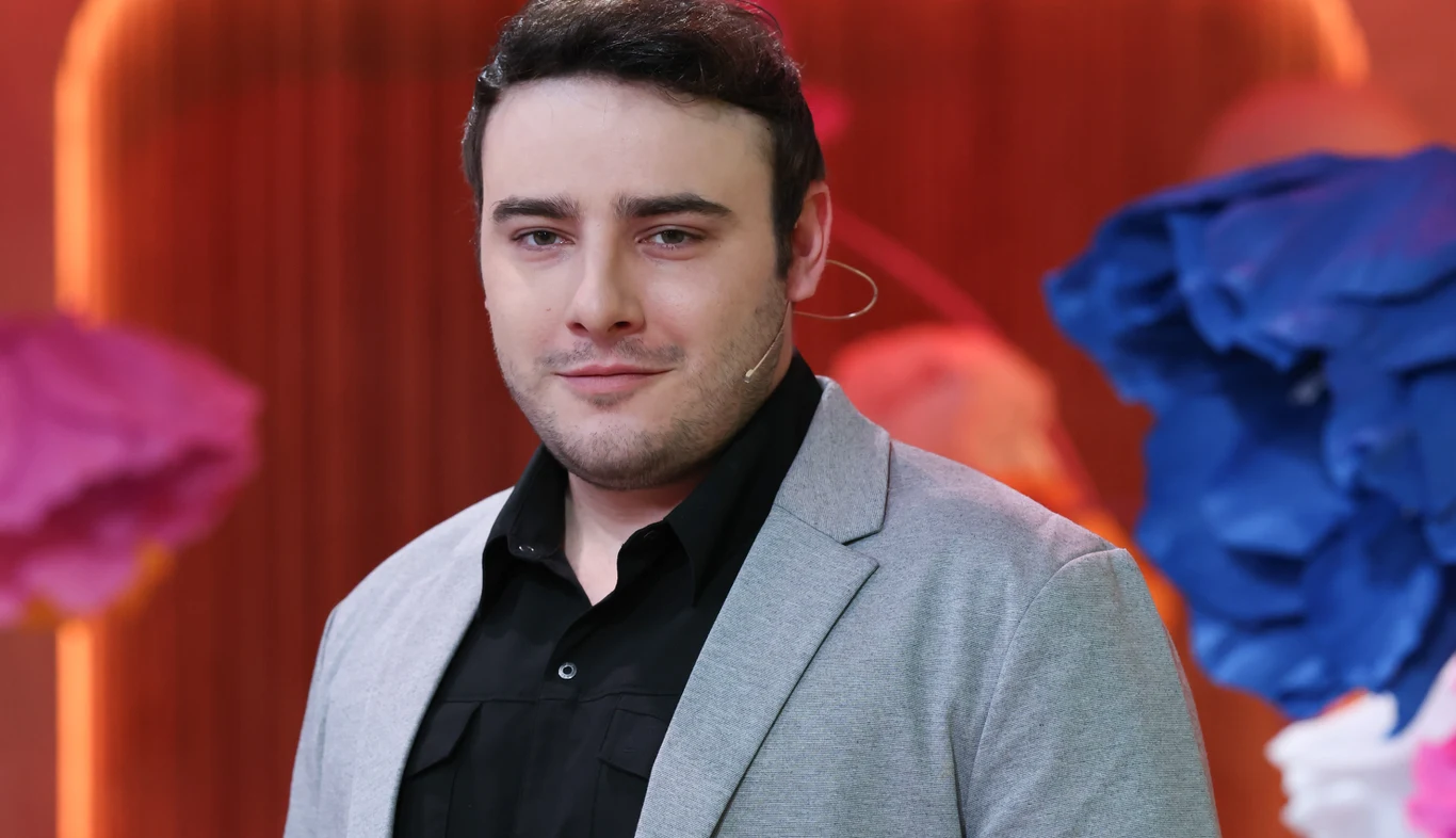 Krystian Ochman wystąpi w show "Twoja Twarz Brzmi Znajomo" Mężczyzna w jasnoszarej marynarce i czarnej koszuli stoi na tle rozmytego pomarańczowego tła z dużymi, kolorowymi dekoracjami w odcieniach niebieskiego i różowego, nosi mikrofon na uchu.