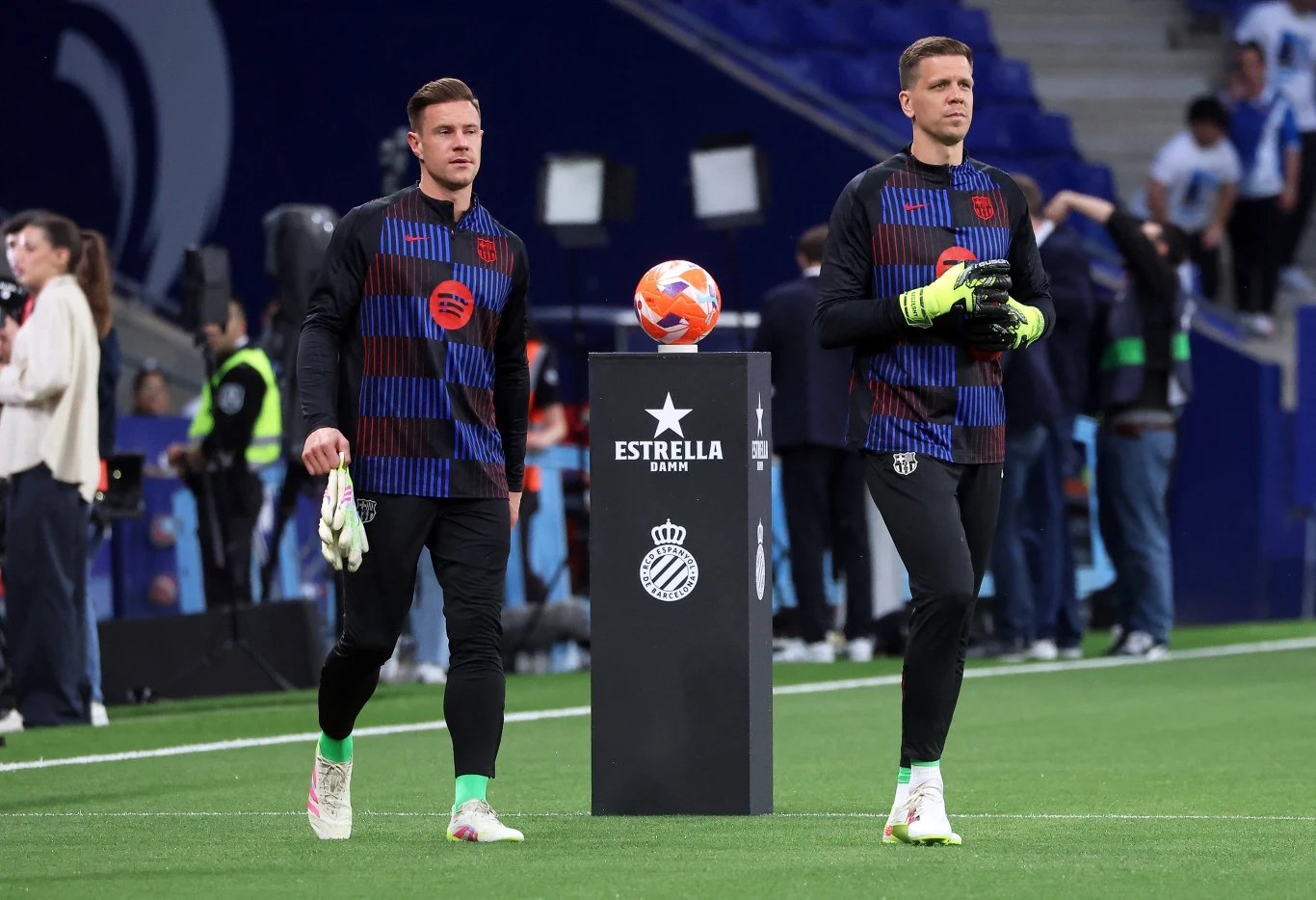 Wojciech Szczęsny i Marc-Andre ter Stegen Dwóch bramkarzy w strojach treningowych piłkarskich klubów stoi na murawie stadionu, obok siebie i naprzeciw symbolicznego podium z logotypem Estrella Damm, w tle widoczni są pozostali zawodnicy oraz fragment trybun.