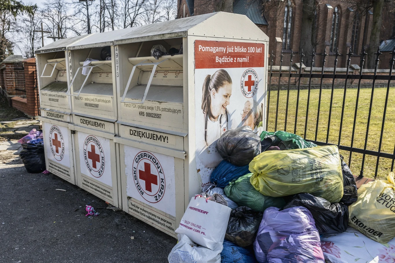 Kontenery PCK znikną z polskich miast Kontenery Polskiego Czerwonego Krzyża przeznaczone do zbiórki odzieży, wokół poukładane worki z ubraniami, na tle ogrodzenia i drzew.