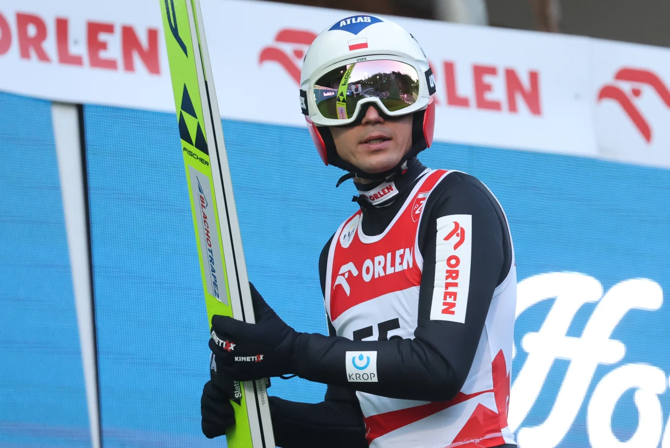Skoczek narciarski w kombinezonie sportowym oraz kasku i goglach stoi z nartą w ręku na tle reklam sponsorskich podczas zawodów.