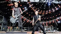 Dwóch muzyków grających na gitarach na dużej scenie koncertowej otoczonej nowoczesnym oświetleniem, w tle perkusista i rozbudowana konstrukcja świetlna.