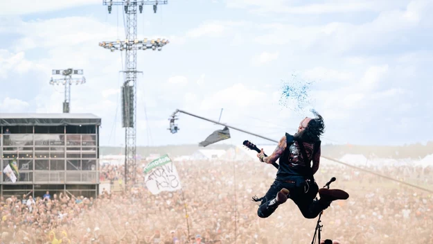 Gitarzysta skacze w powietrzu na scenie podczas koncertu, trzymając gitarę, z kropelkami wody unoszącymi się wokół jego głowy, w tle tłum widzów oraz scena z techniczną infrastrukturą.
