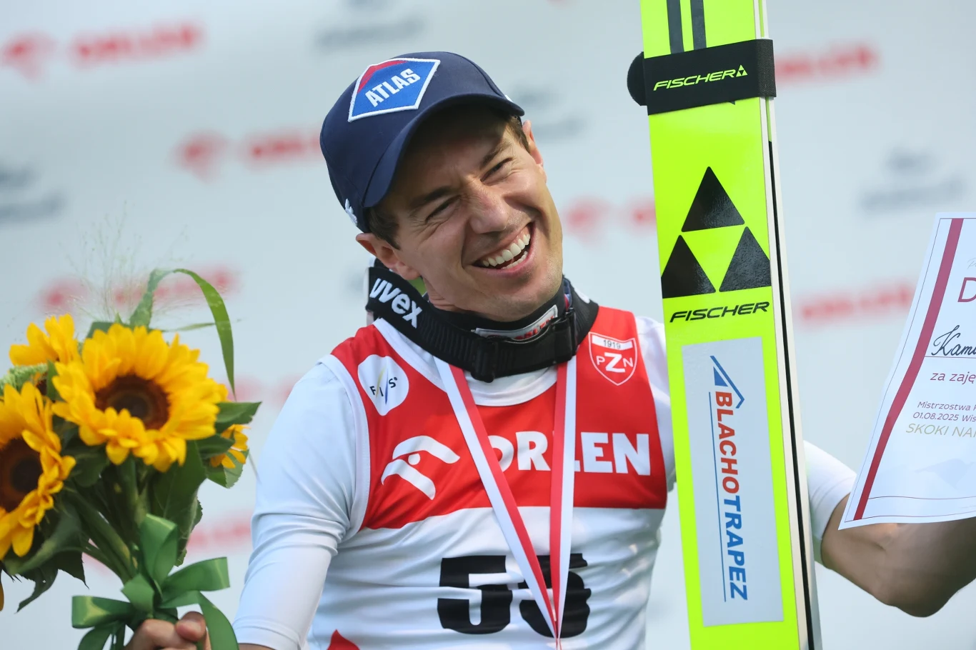Sportowiec w sportowym stroju z medalem na szyi, uśmiechnięty trzyma bukiet słoneczników oraz narty, w tle rozmyte logo sponsorów.
