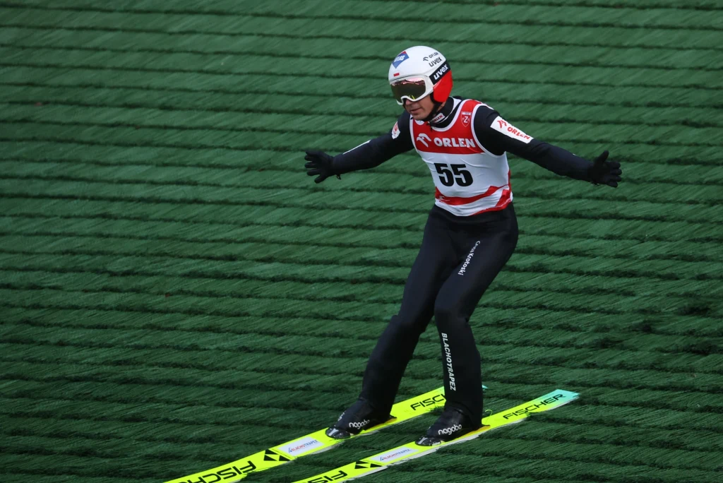 Kamil Stoch Skoczek narciarski w pełnym ekwipunku ląduje po skoku na zielonej, sztucznej macie, z rozłożonymi ramionami w celu utrzymania równowagi, widoczny numer startowy 55 na kombinezonie.