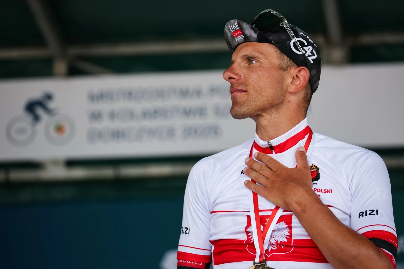Kolarz w biało-czerwonym stroju z orłem na piersi stoi przed tablicą z napisem dotyczącym mistrzostw Polski, na szyi ma medal, a ręką trzyma się za pierś.