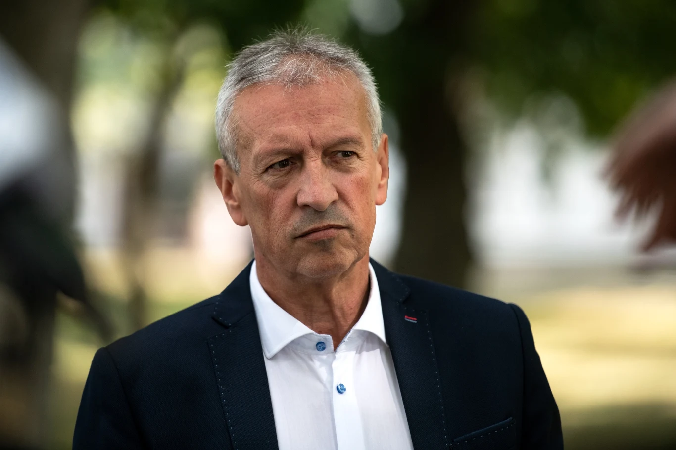 Jacek Dobrzyński Starszy mężczyzna w garniturze i białej koszuli stoi na świeżym powietrzu, w tle rozmyte drzewa i zielona przestrzeń, twarz skupiona i poważna.